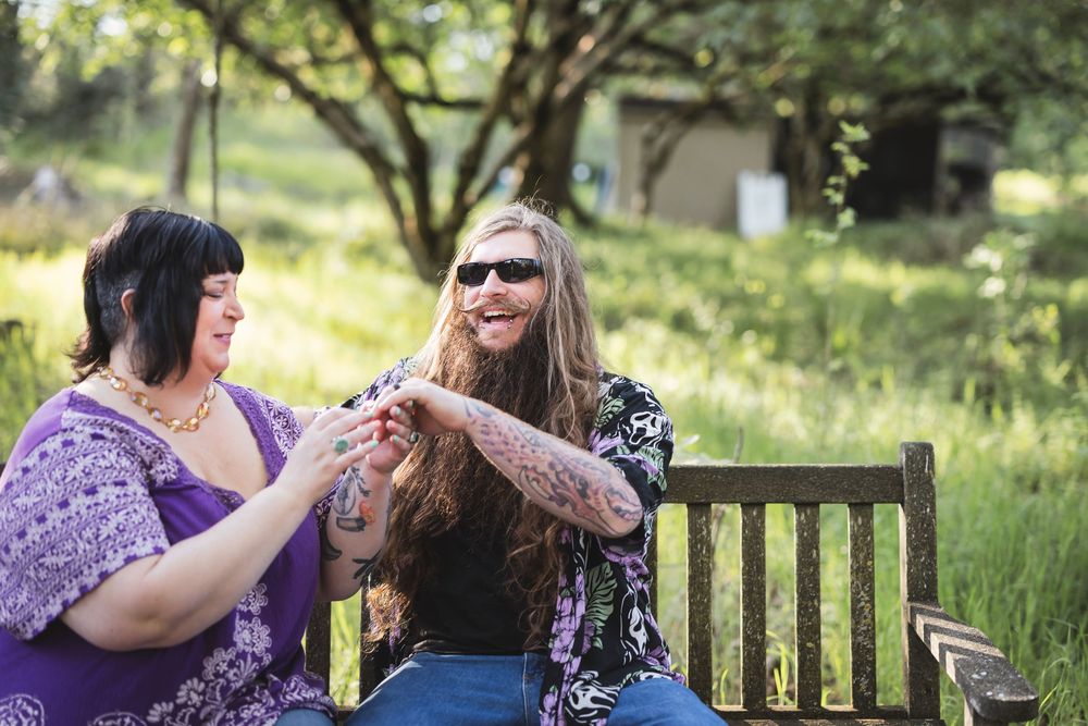 Two friends share a laugh while sitting together on a wooden bench in a sunny garden setting.