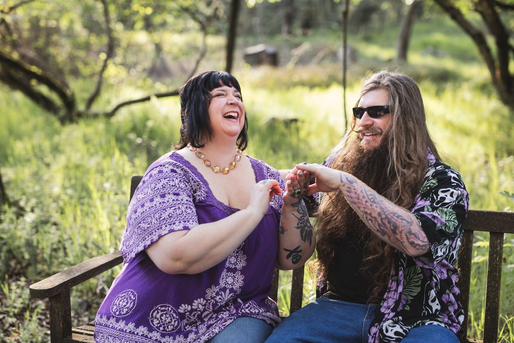 Two friends share a joyful moment while sitting together on a wooden bench in a sunny outdoor setting.