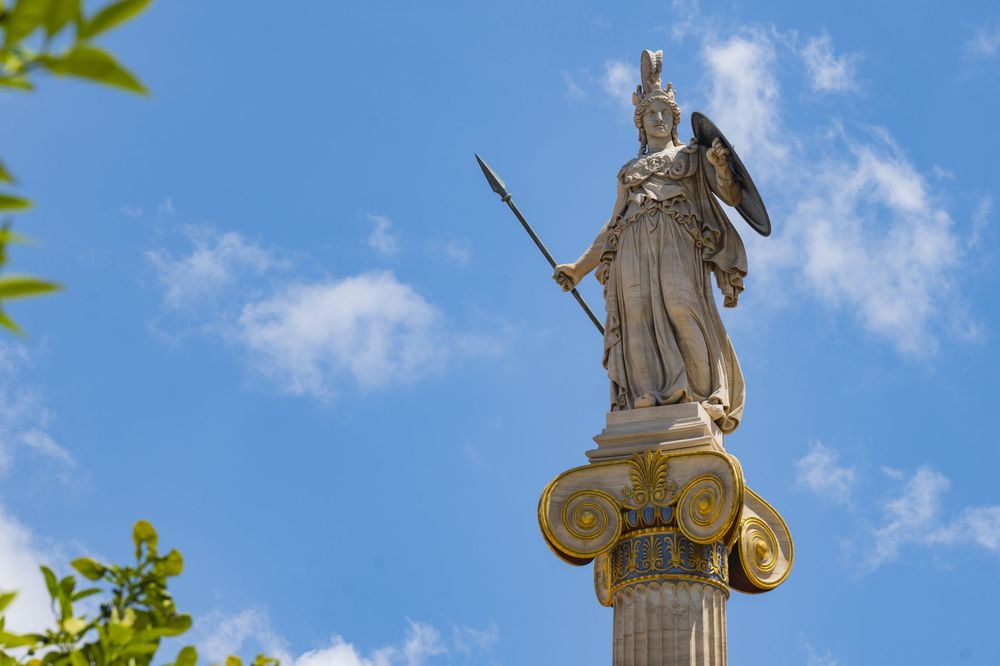 Ornate marble statue of a warrior figure stands atop a tall pillar against a bright blue sky with scattered clouds.