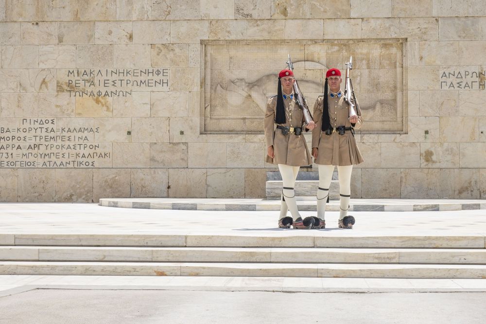 Two guards in traditional uniforms stand at attention during the changing of the guard ceremony at a stone monument.