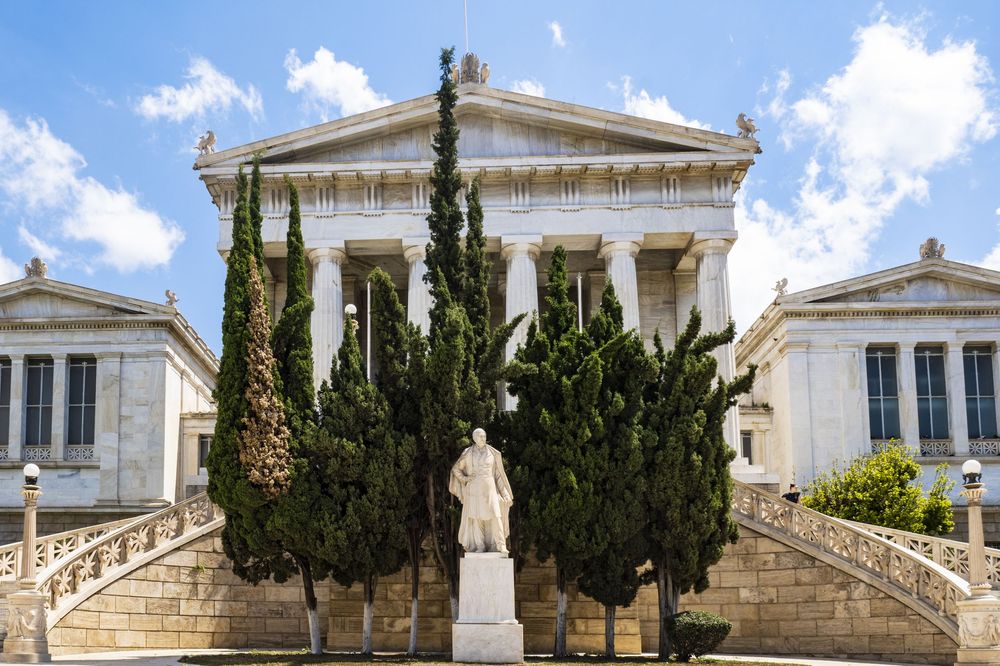 Neoclassical buildings with white columns and a central statue stand behind tall cypress trees at the National Library of Greece.