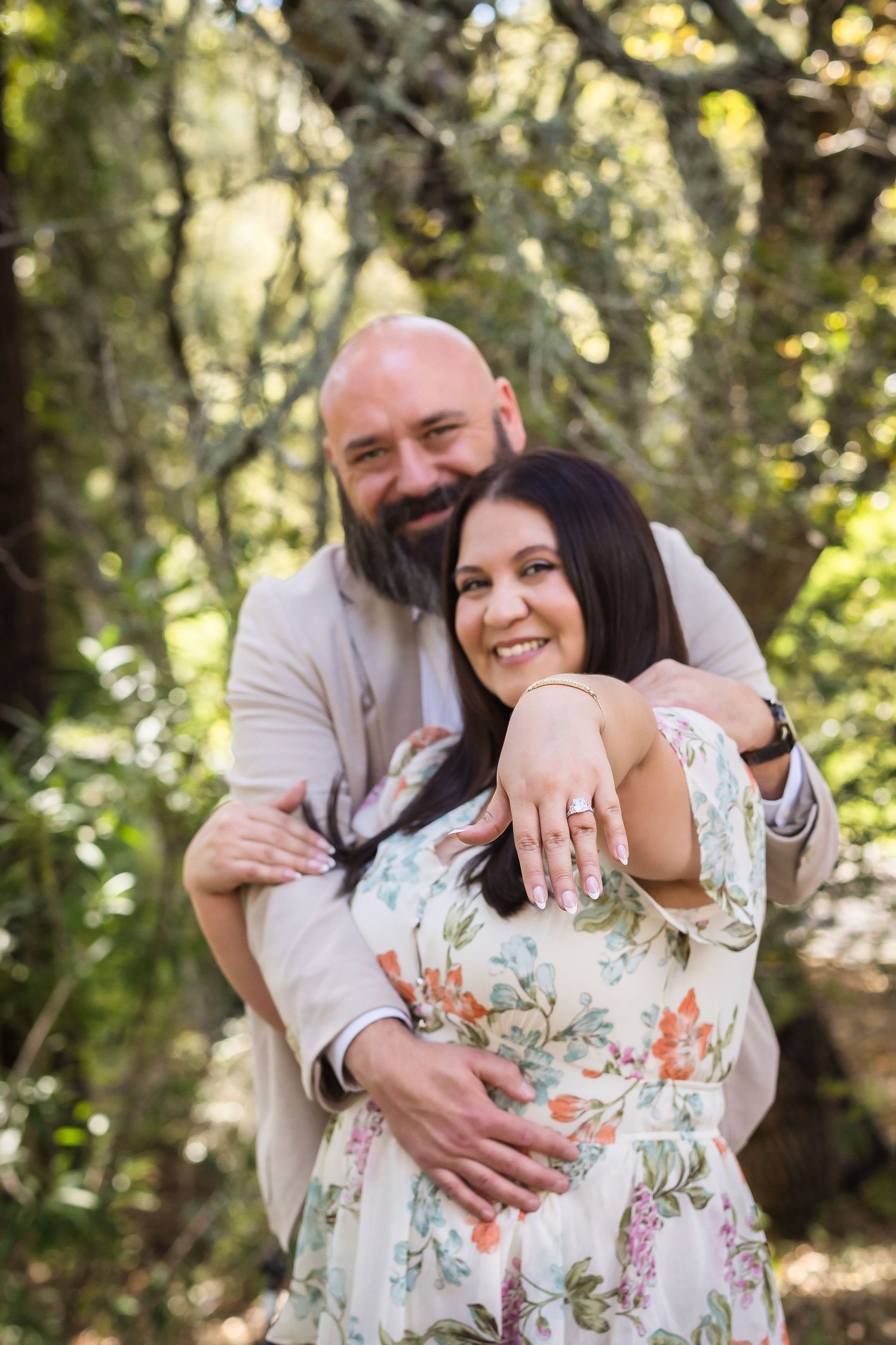 A couple embraces outdoors in a natural setting with sunlit trees in the background.
