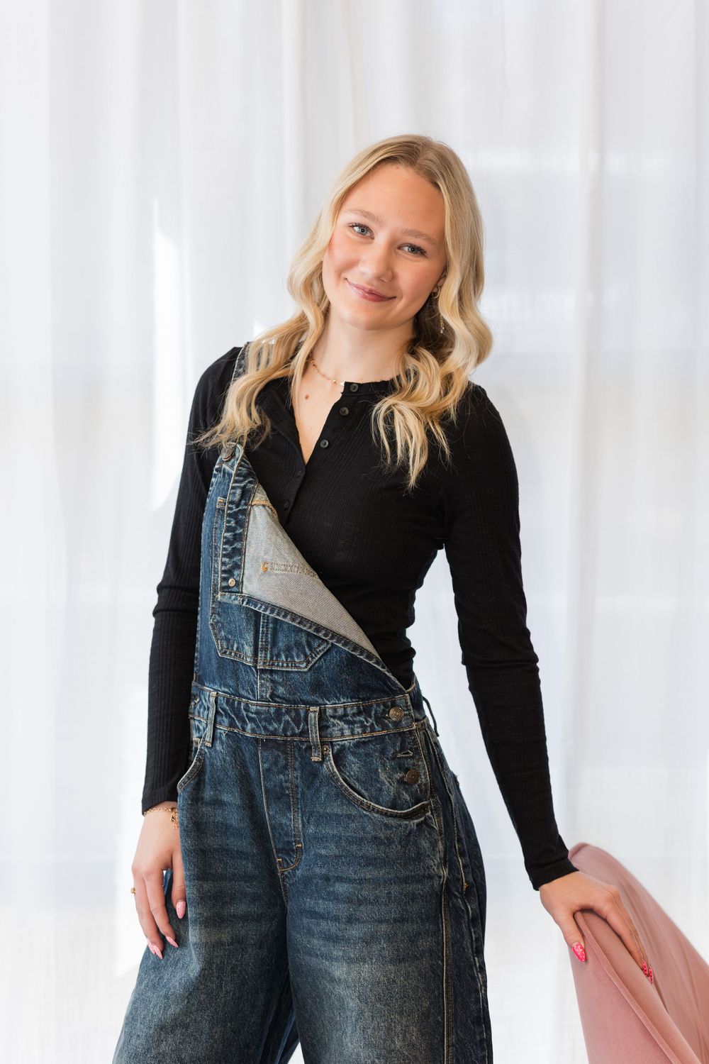 Woman in black long sleeve top and denim overalls poses against white backdrop in casual style photoshoot.