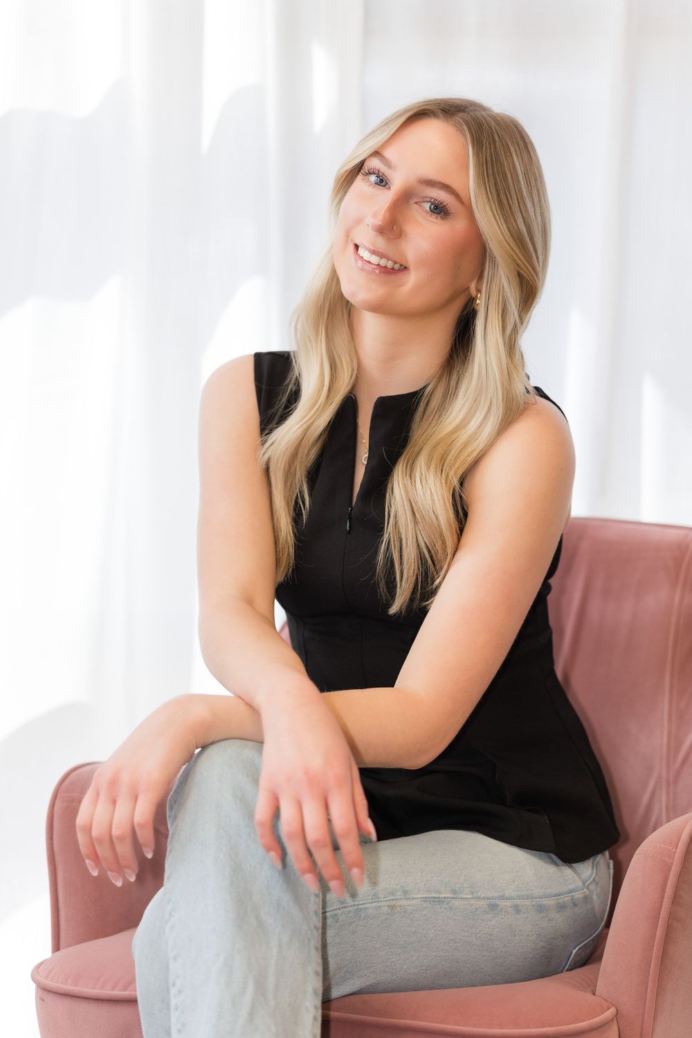 Woman in black sleeveless top poses casually on pink chair against white backdrop.