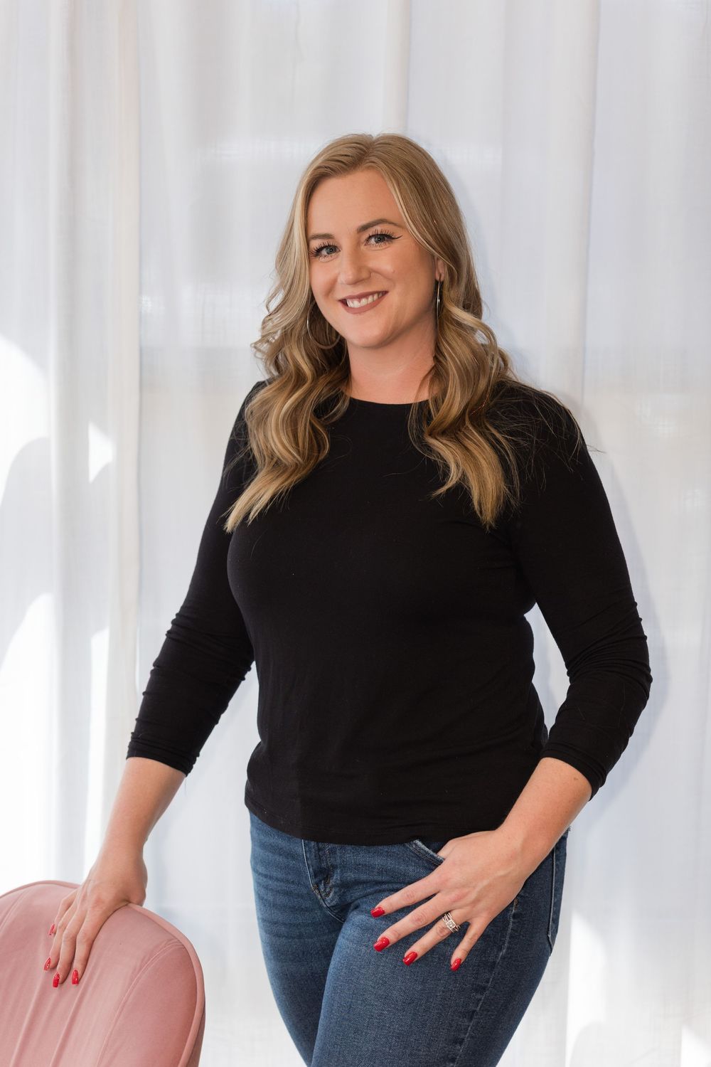 Woman in black long sleeve top and jeans poses against white backdrop with confident smile.