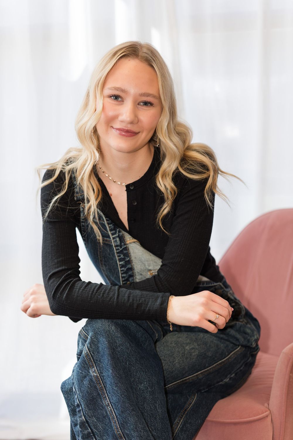 Casual portrait of someone wearing a black long sleeve top and denim overalls while sitting on a pink chair.