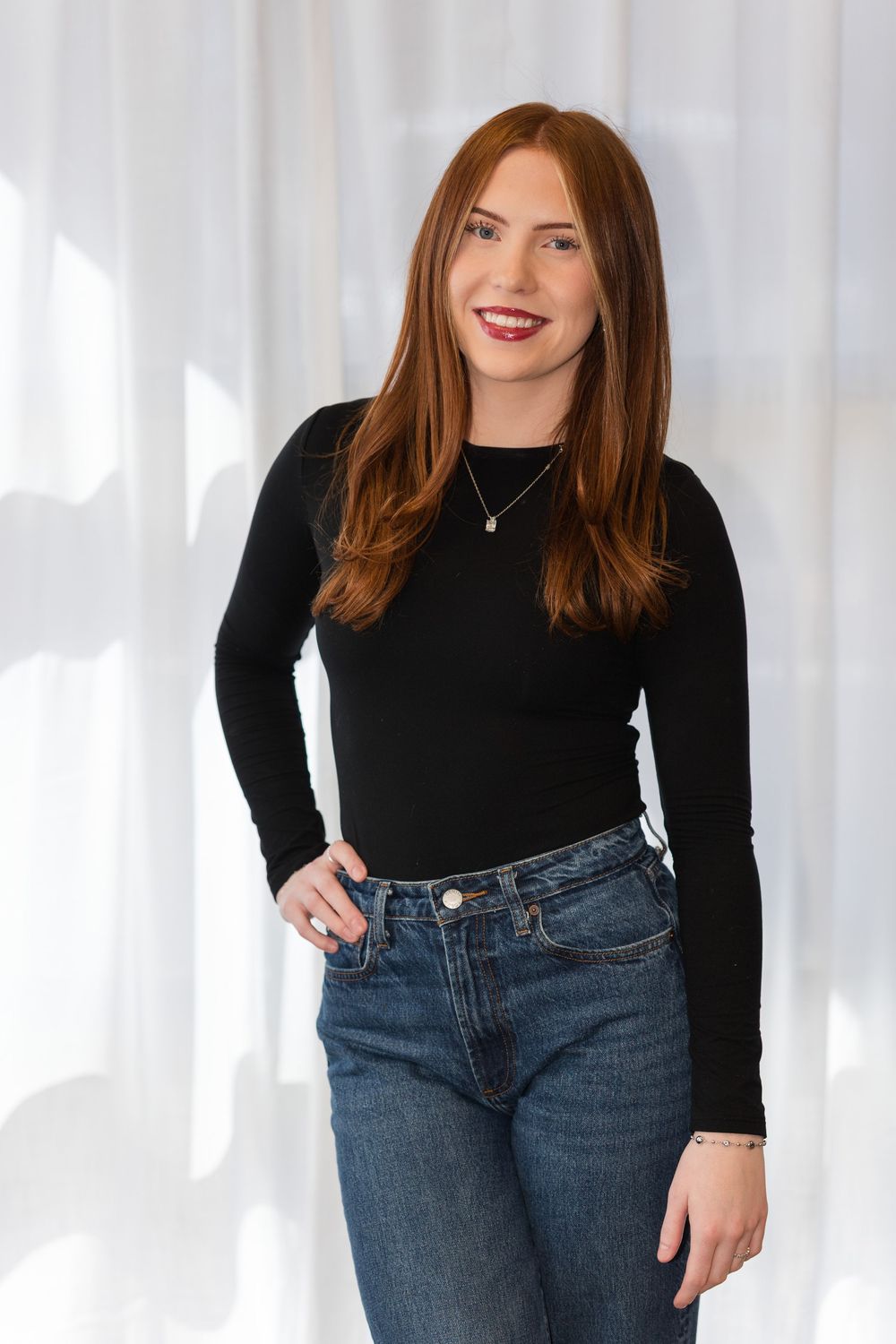 Model wearing casual black top and blue jeans poses against white curtain background.