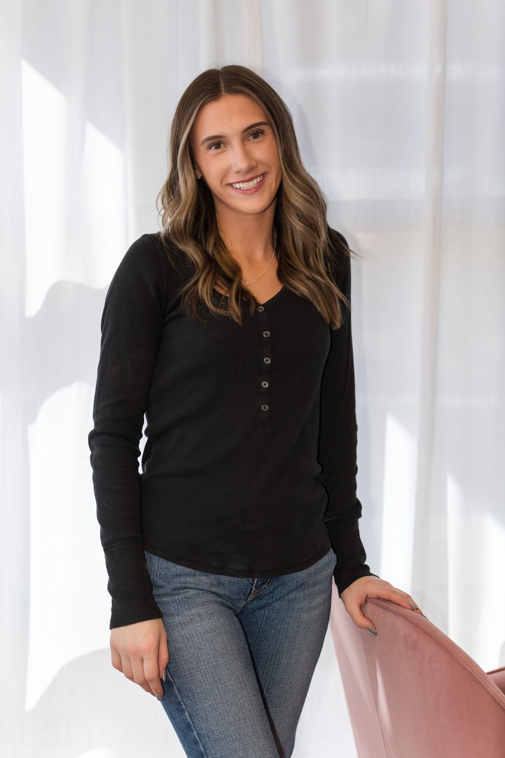 Woman in black sweater and jeans leans against pink chair in studio setting.