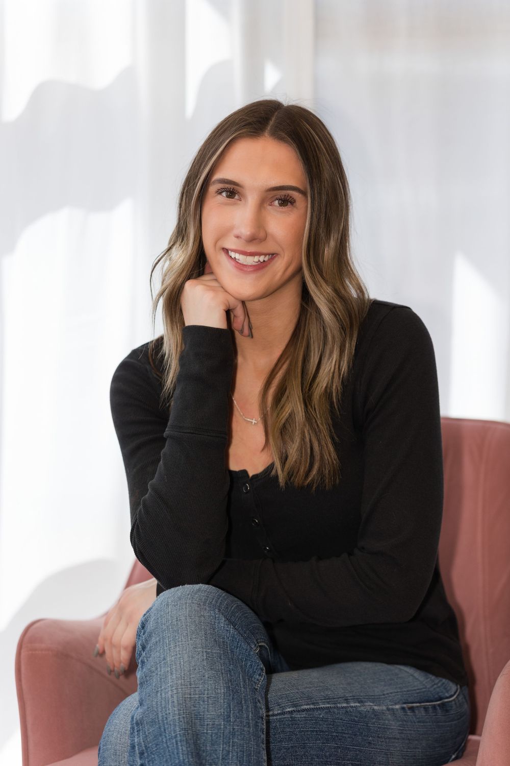Professional portrait of someone in a black sweater and jeans sitting casually on a pink chair against a white backdrop.