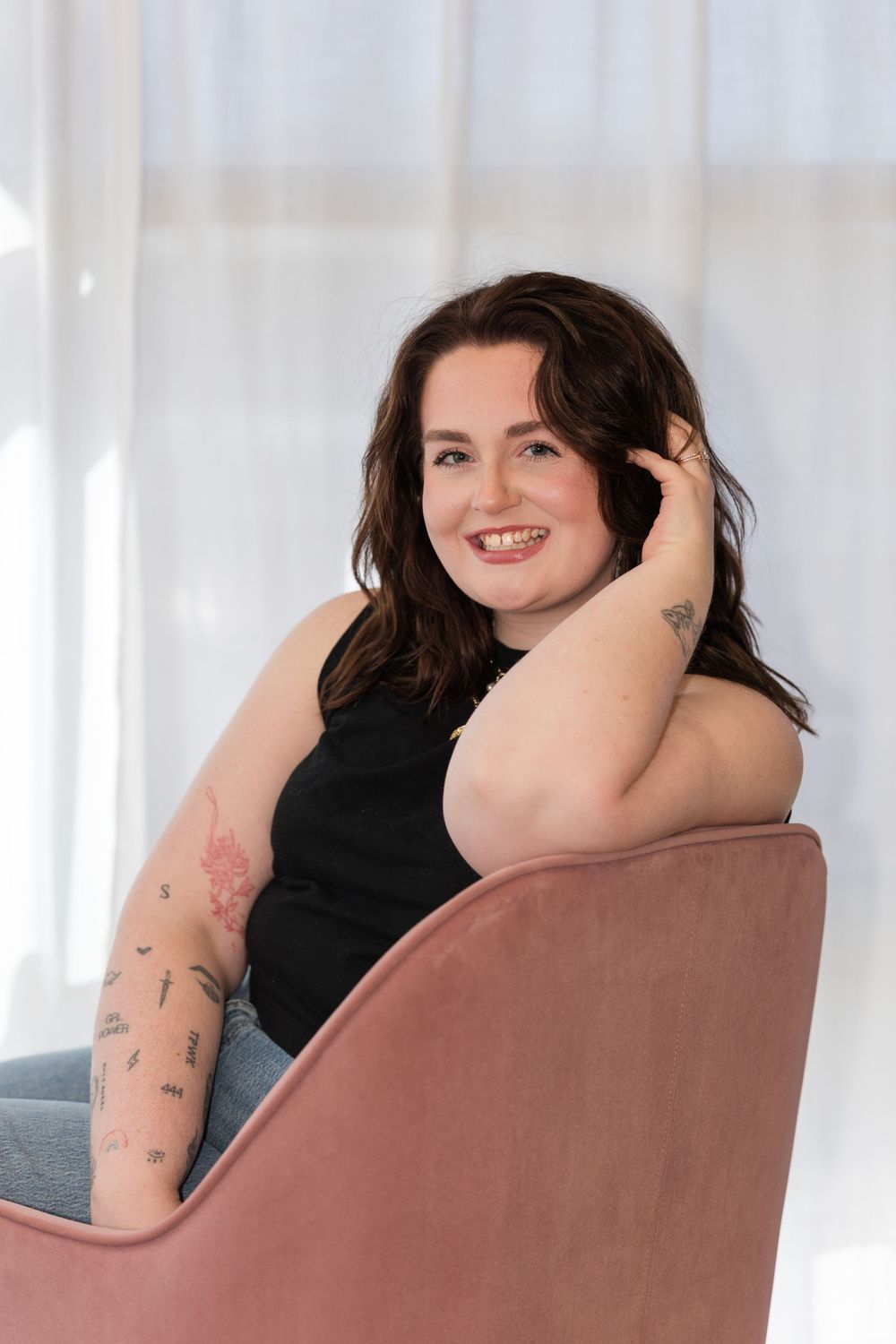 Woman relaxes in pink chair wearing dark top and jeans with visible arm tattoos.