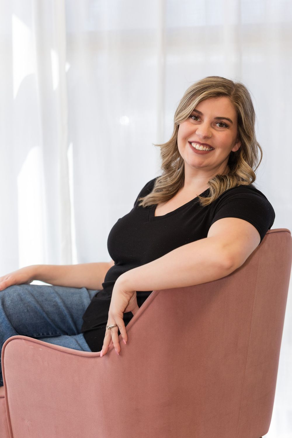 A person in a black top and jeans sits casually in a pink velvet armchair by a bright window.