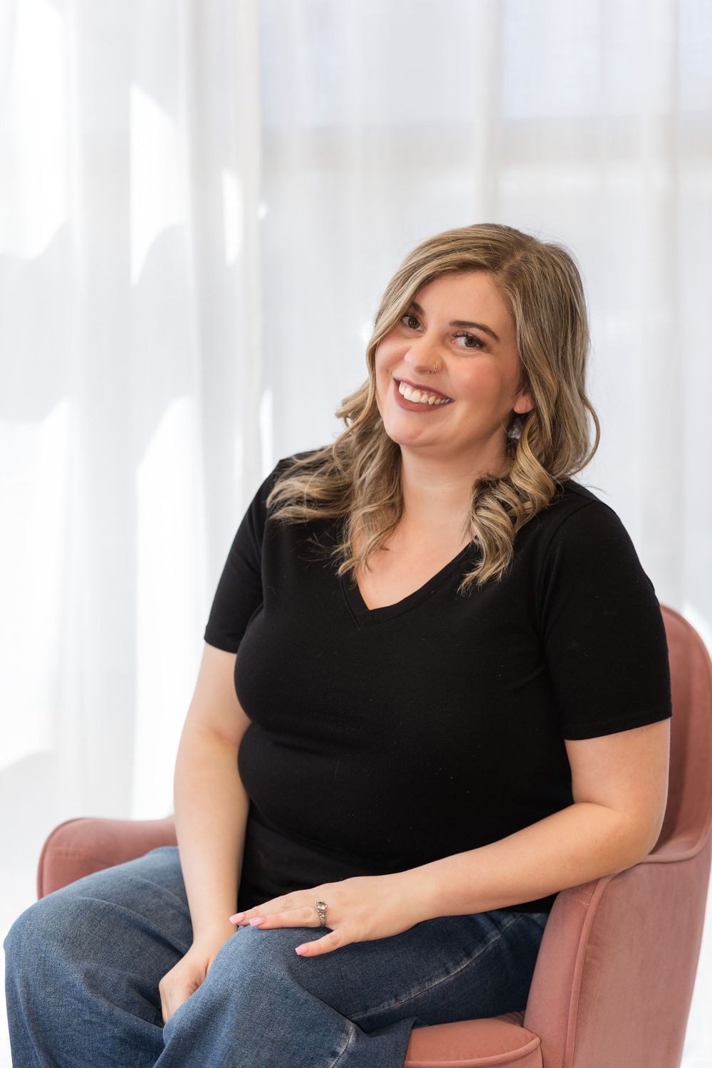 Woman in black shirt smiles and laughs while sitting on pink chair in bright room with natural lighting.