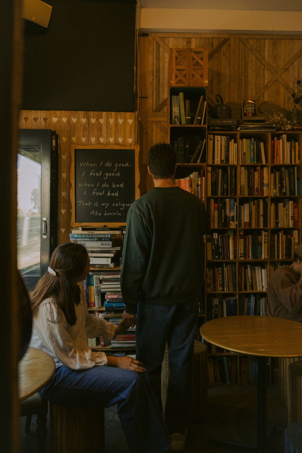 Two figures browse books together in a cozy library with wooden shelves and warm lighting.