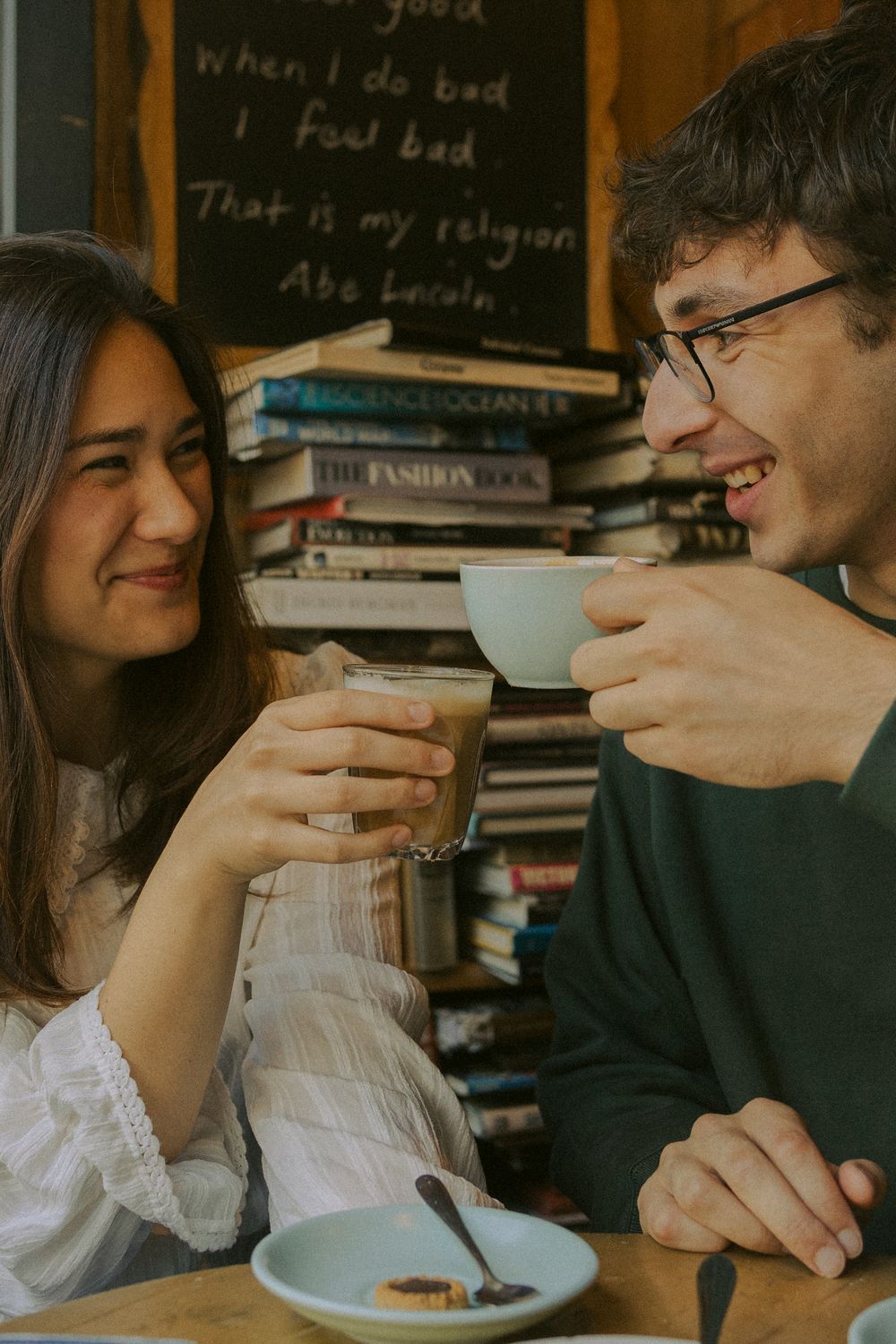 Couple enjoying coffee and conversation at a cozy cafe with bookshelves and chalkboard writing in the background.