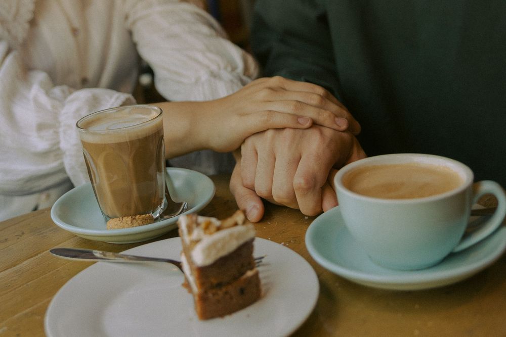Hands holding various coffee drinks in white ceramic cups on a wooden table with natural lighting.