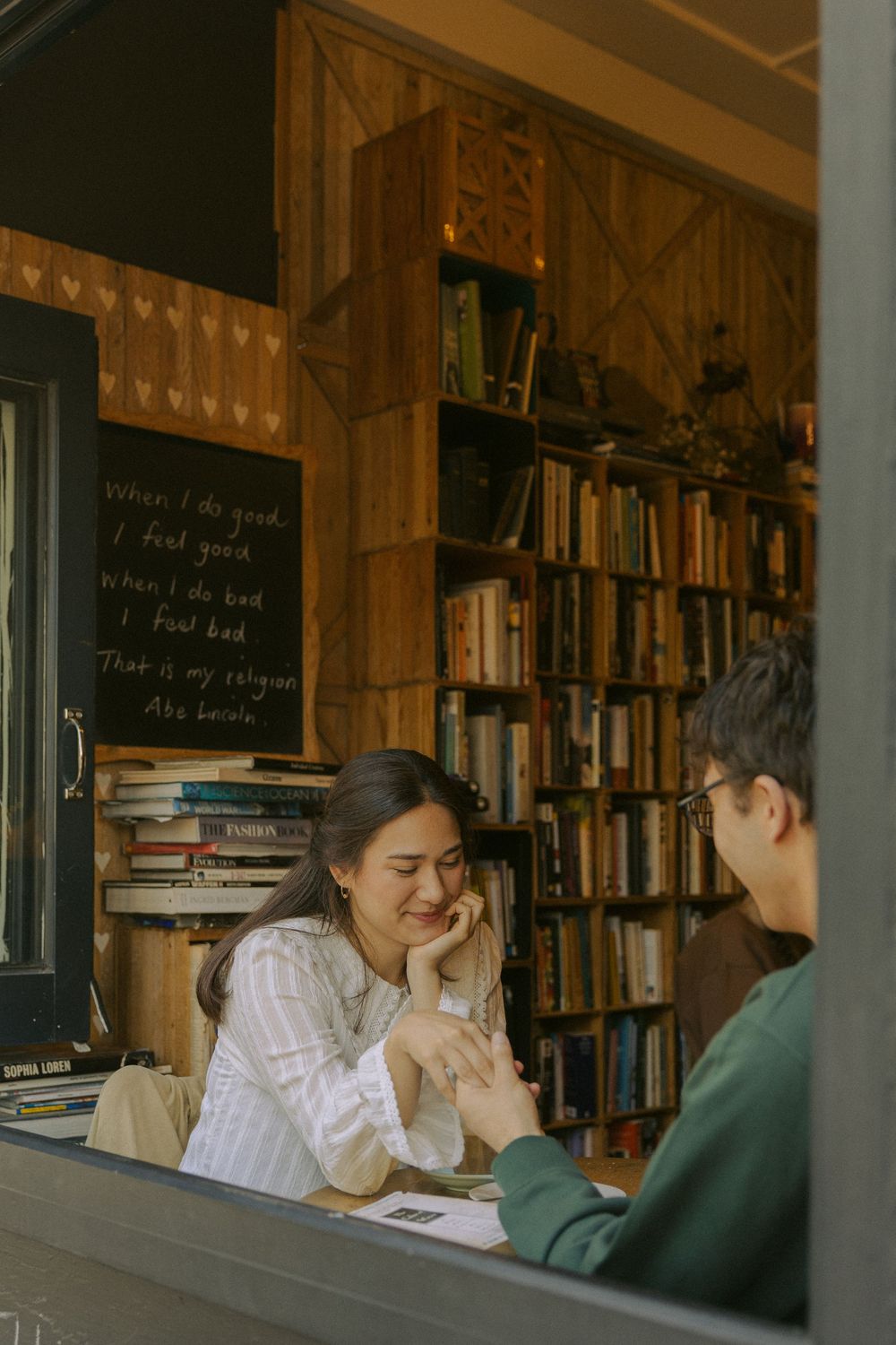 Two people share a cozy moment in a library-like setting with wooden bookshelves and a chalkboard in the background.