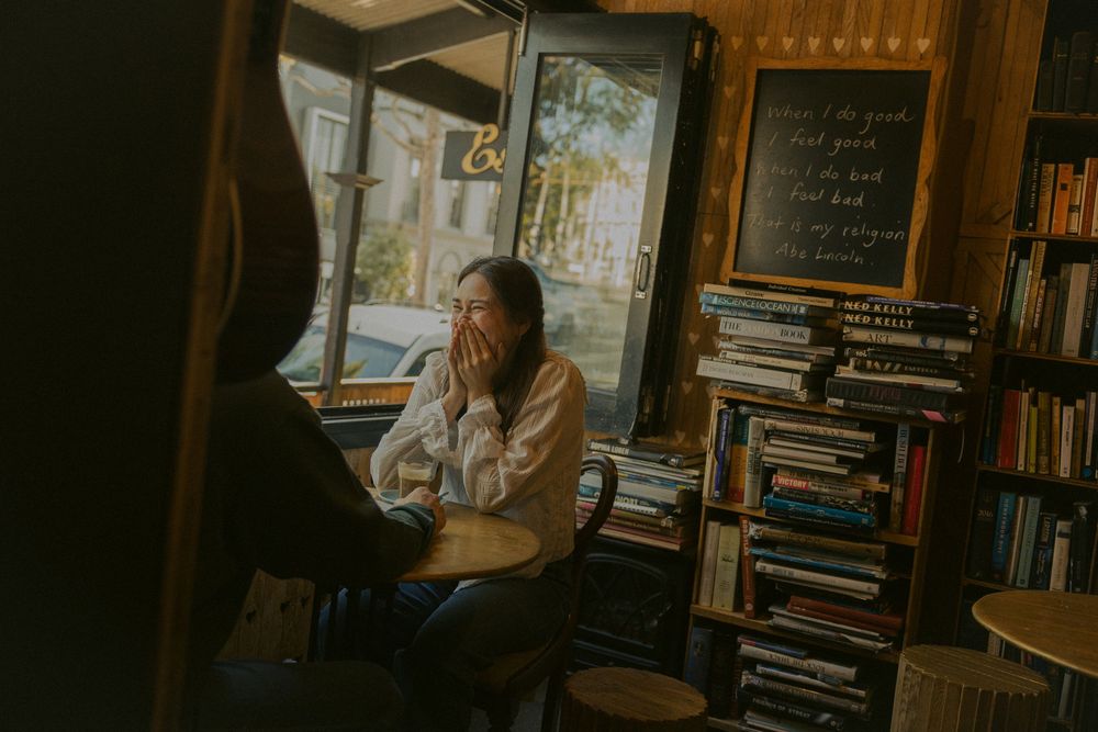 Person sits thoughtfully in cozy library surrounded by bookshelves and natural light streaming through window.