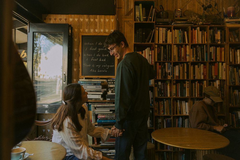 Two people having an intimate conversation in a cozy library with bookshelves and natural light streaming through a window.