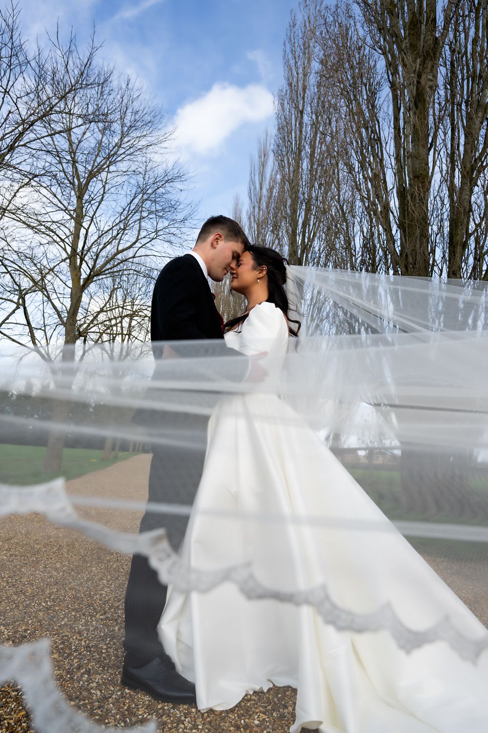 A romantic wedding couple embraces outdoors while a flowing white veil creates an ethereal effect against bare winter trees.