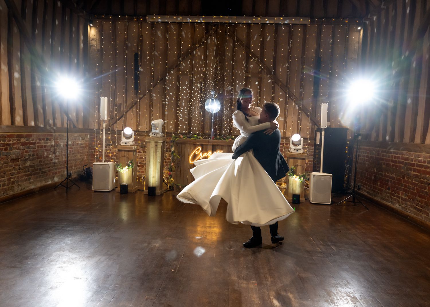 A rustic barn wedding venue with exposed brick walls and twinkling lights illuminating the wooden dance floor.