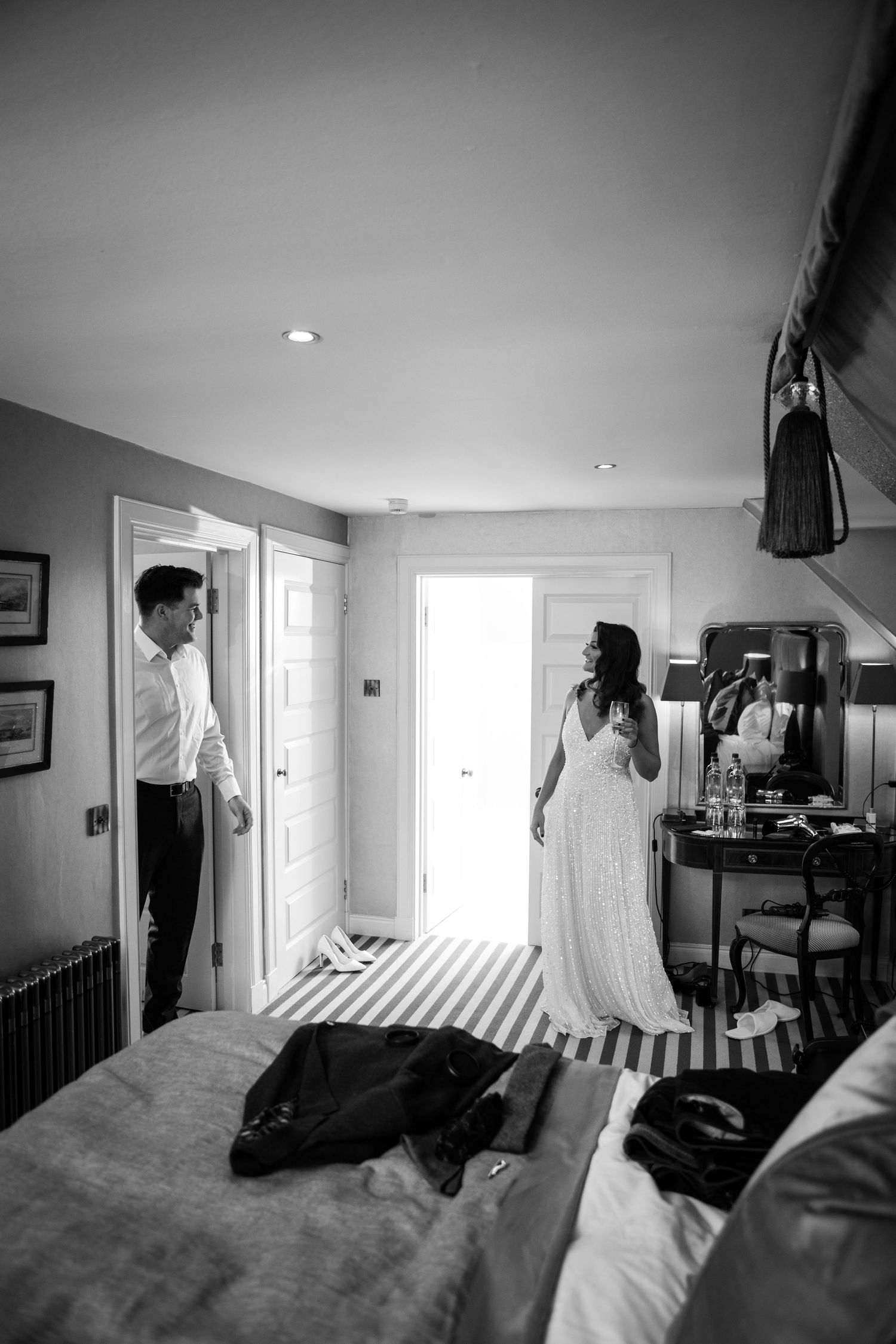 Black and white wedding day photograph showing a couple in a bedroom with sunlight streaming through windows.