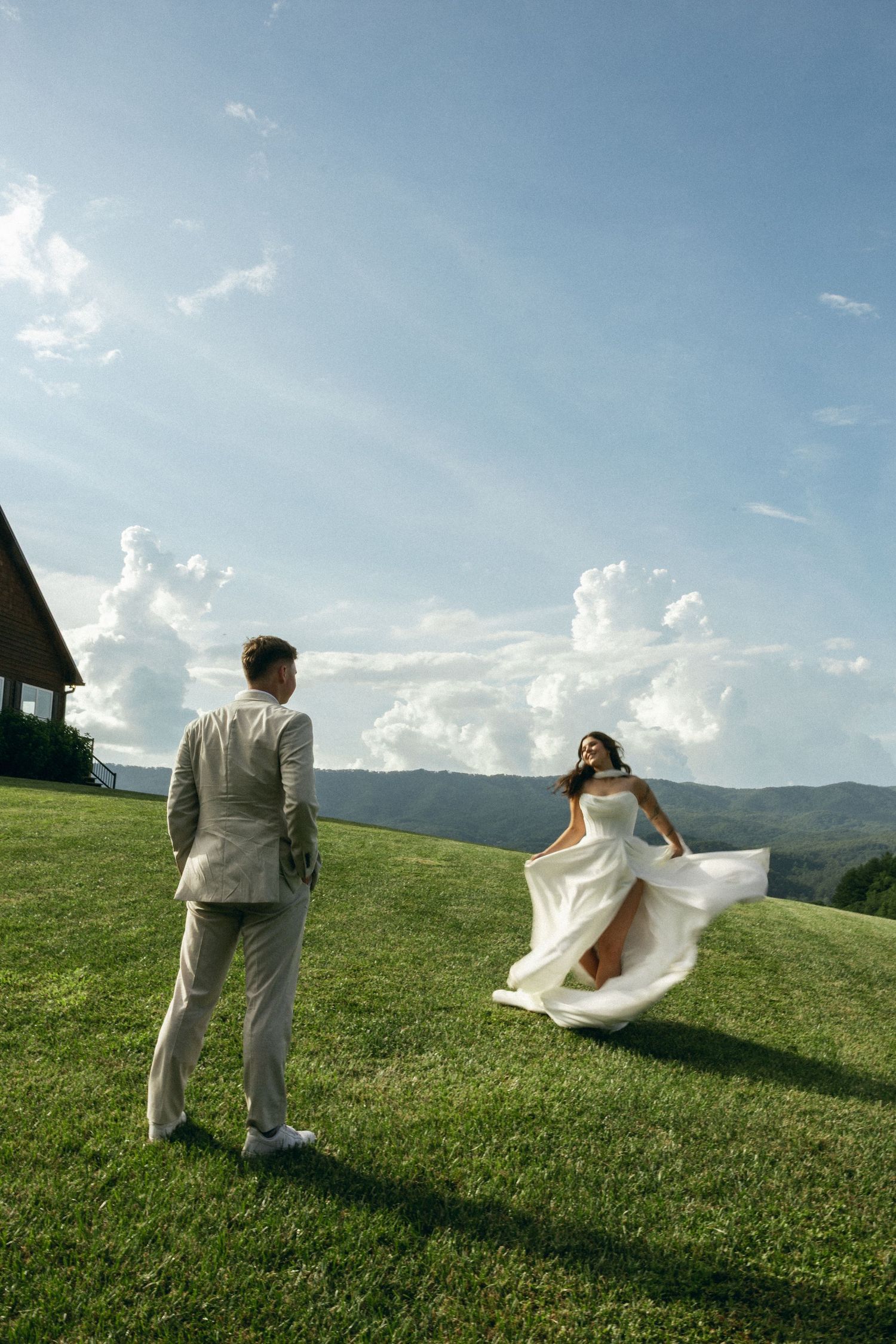 A couple in wedding attire stands on a hilltop meadow with mountains and dramatic clouds in the background.Micro wedding, elopement, intenti