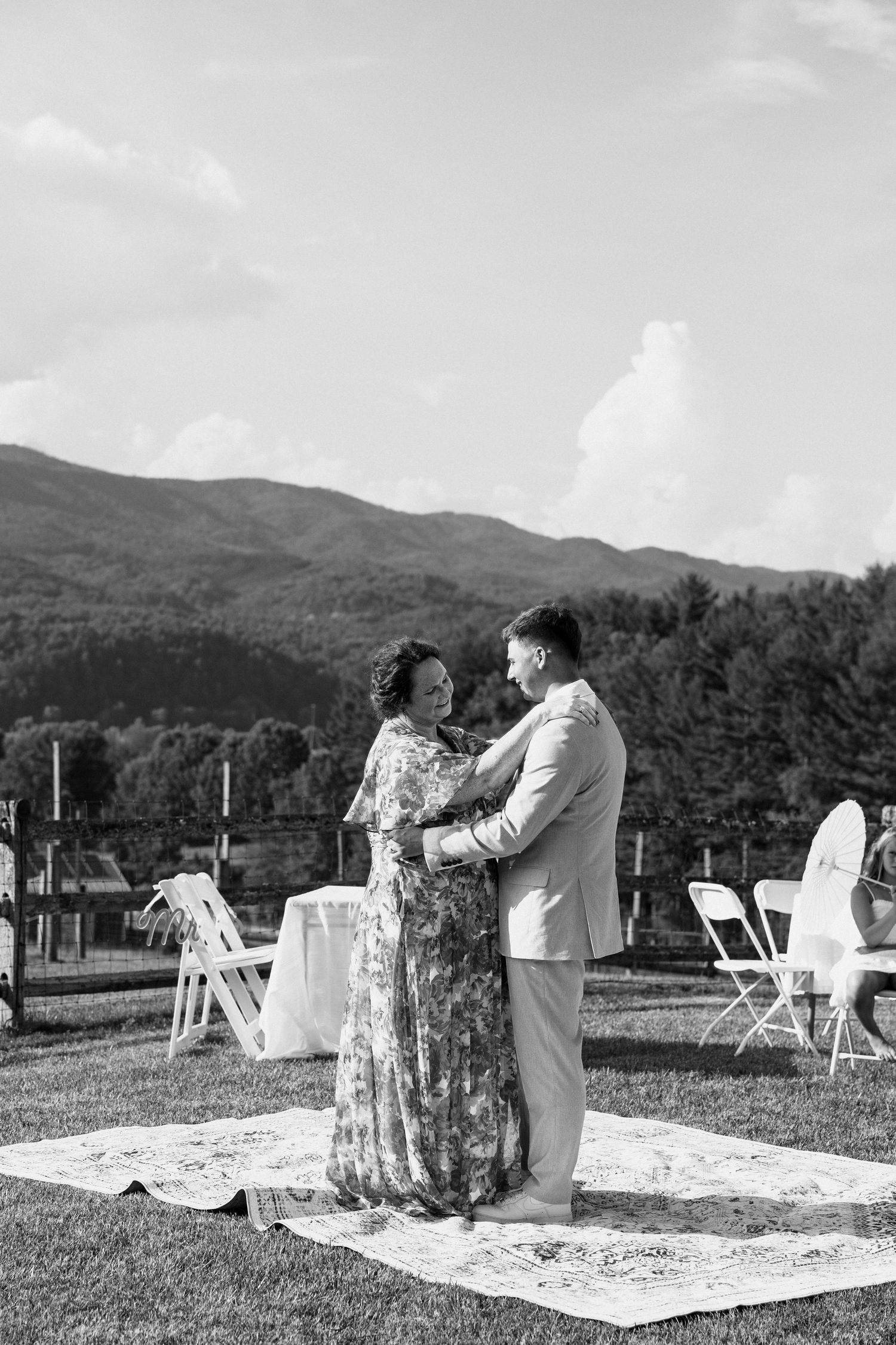 People gather on blankets for an outdoor event with white chairs and mountain views in the background.