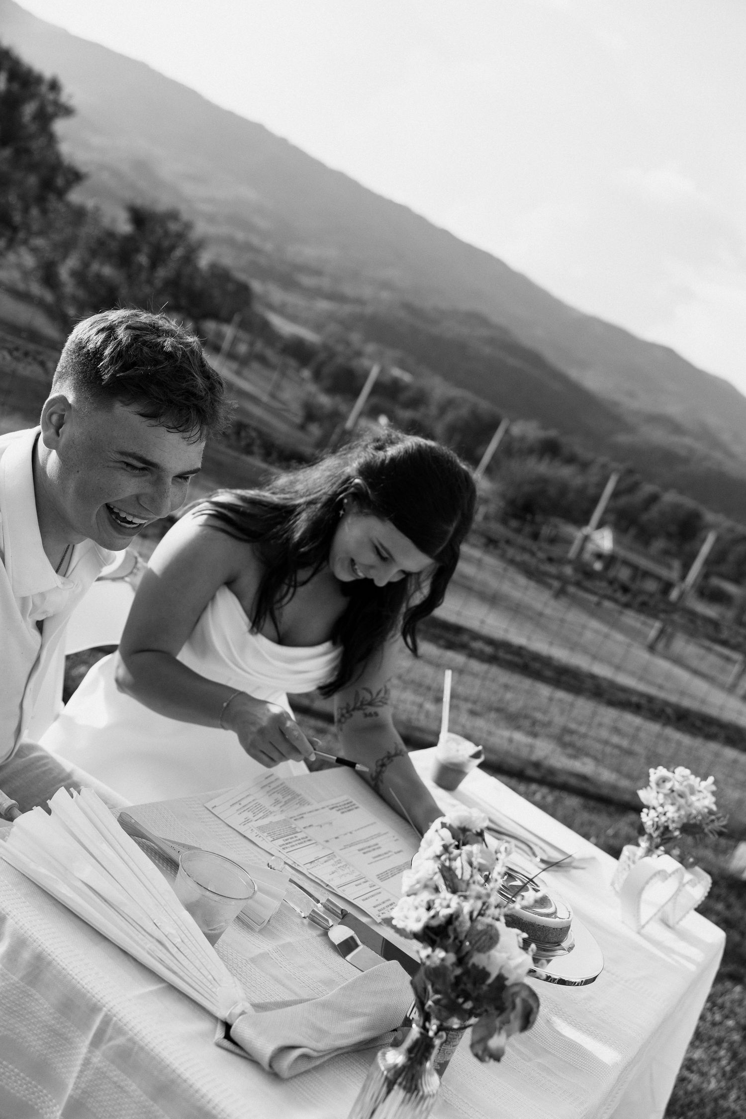 A couple in white attire share a joyful moment while signing papers at an outdoor table with mountains in the background. Micro wedding, elo