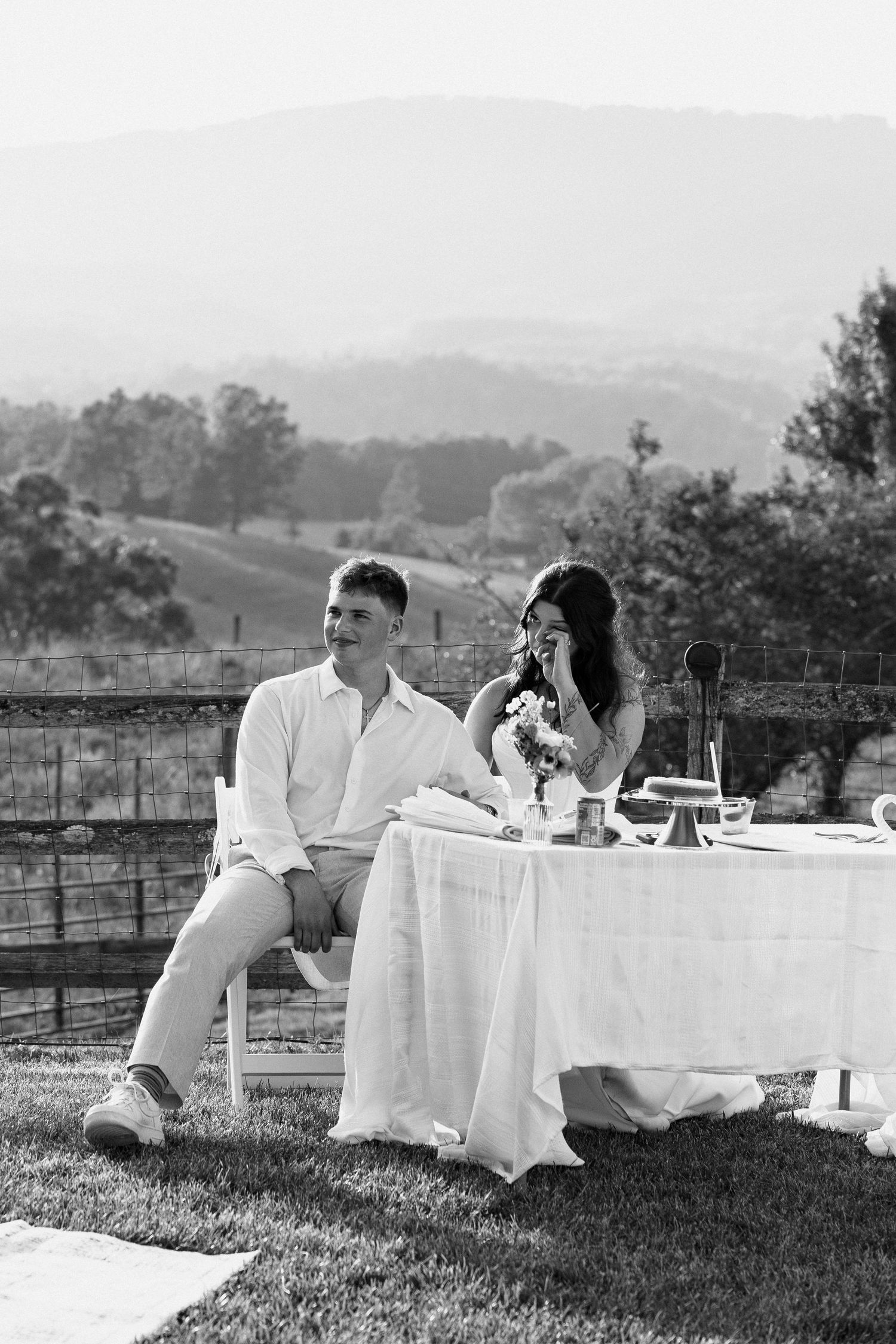 A couple in white attire shares an intimate moment at an outdoor table setting with rolling hills in the background. Micro wedding, elopemen