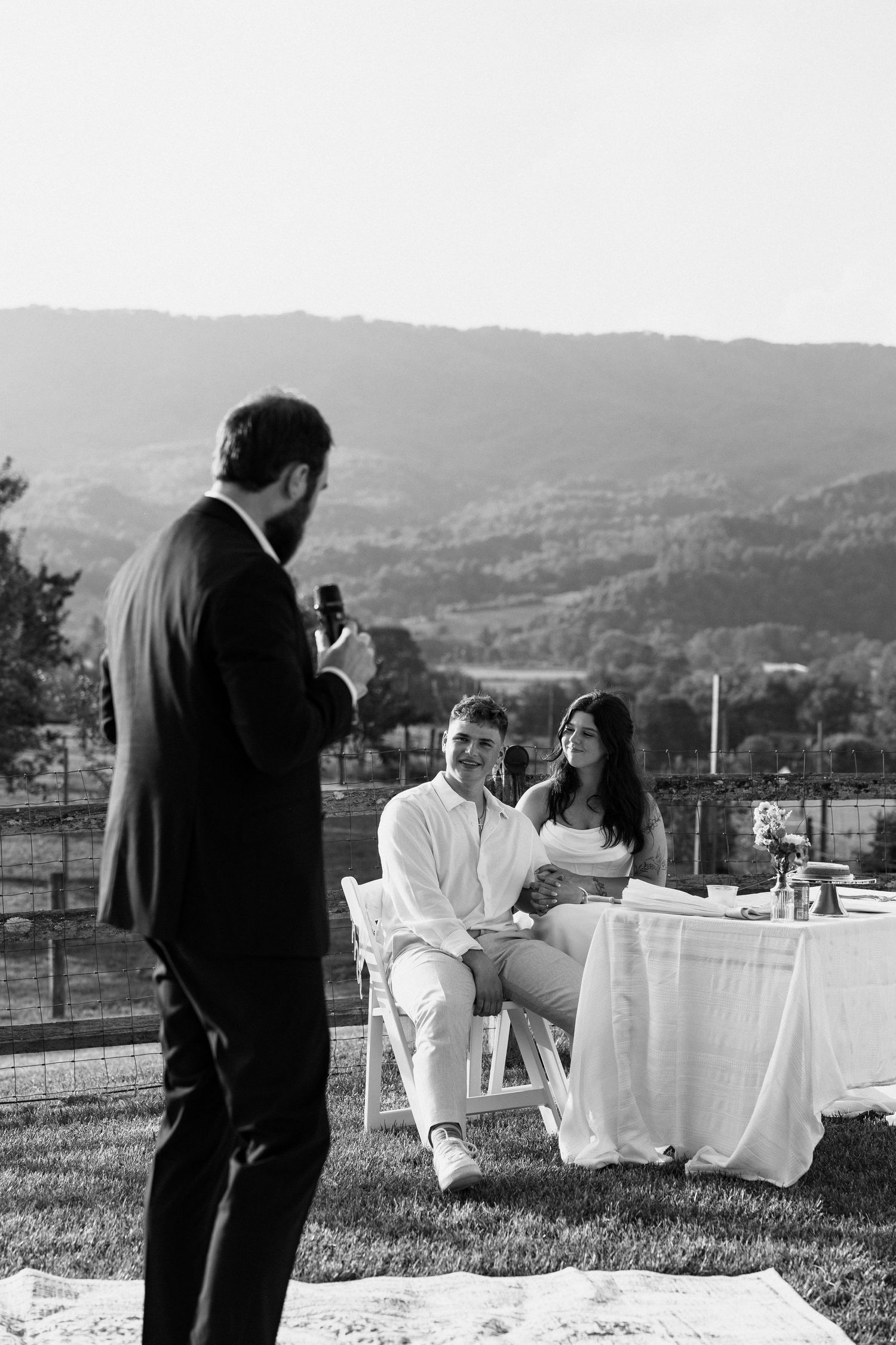 A black and white outdoor photoshoot with people seated on chairs against a scenic hillside backdrop. Micro wedding, elopement, intentional