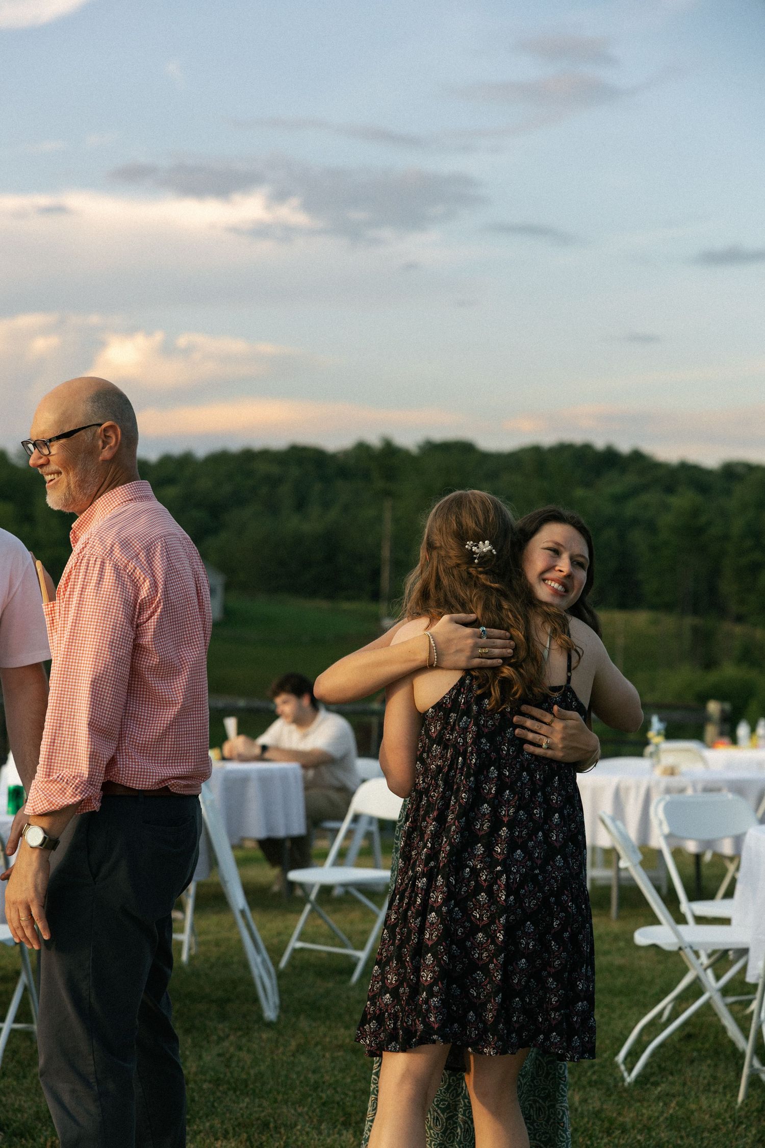 Wedding guests dance together at an outdoor reception during sunset with white tables in the background.