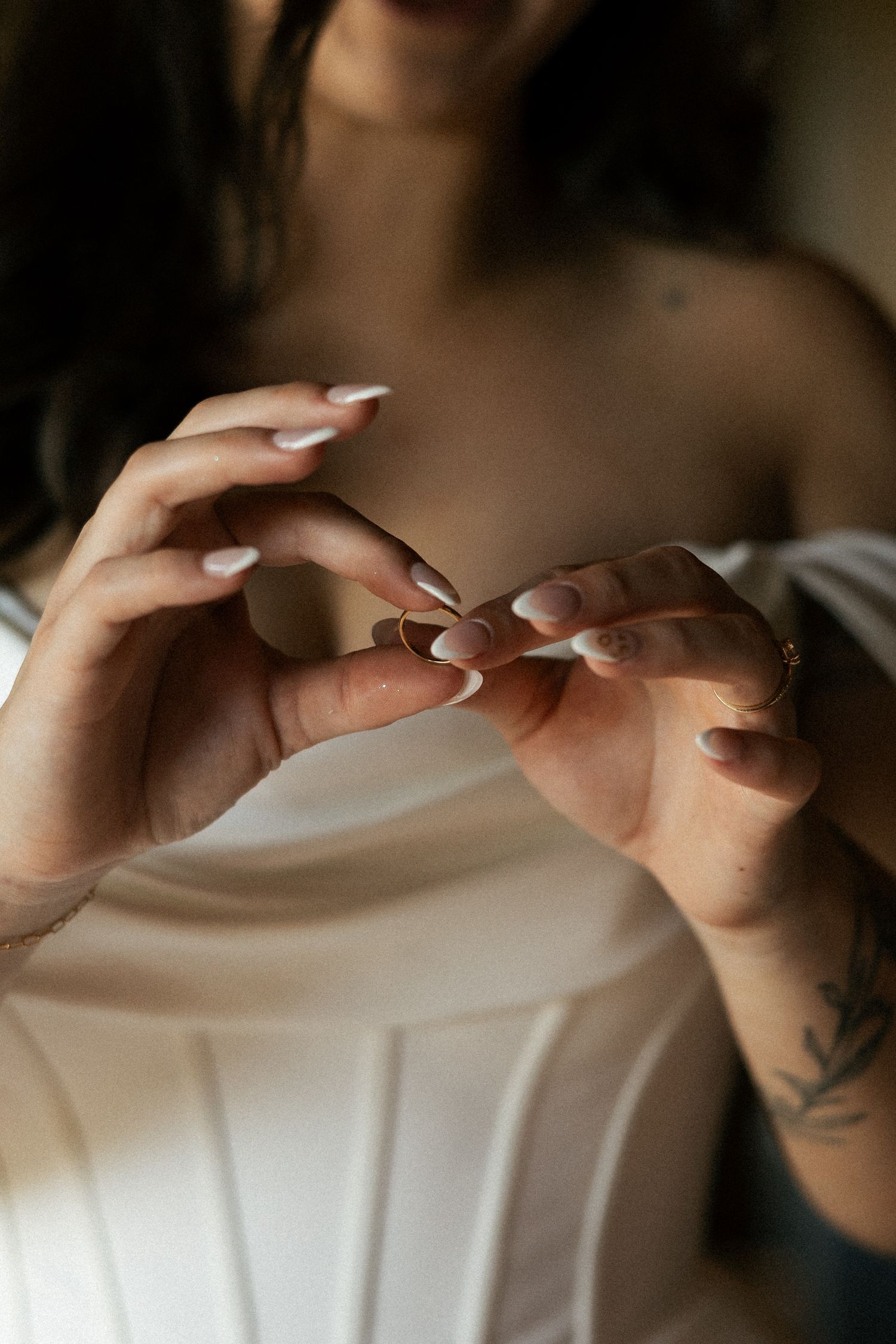 Hands with white manicured nails and tattoos fastening delicate jewelry against white fabric.Micro wedding, elopement, intentional celebrati