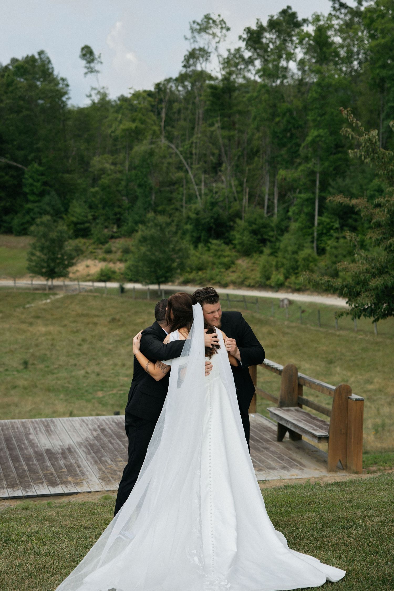 Three people gather around a flowing white wedding dress on a wooden deck surrounded by lush green trees.