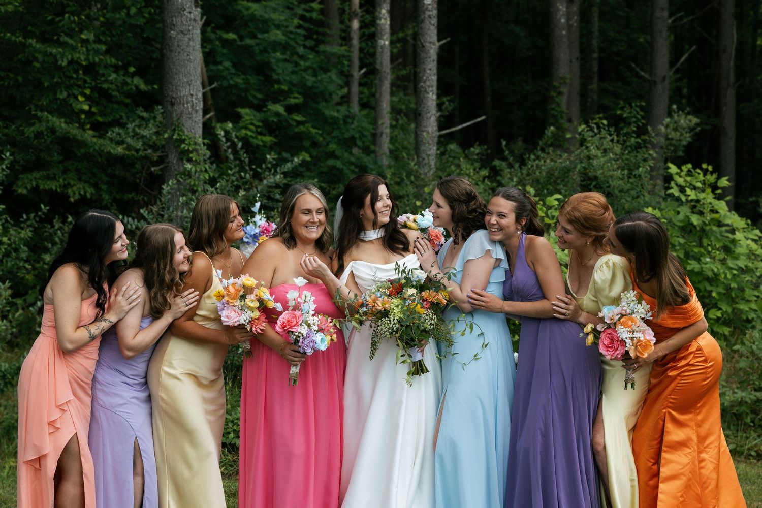 Wedding party wearing colorful pastel dresses stands in line against green forest backdrop.