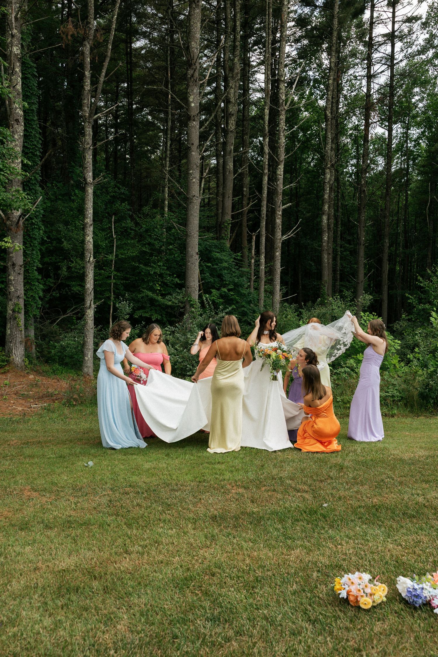A group in pastel bridesmaid dresses help arrange a wedding dress in a wooded outdoor setting.