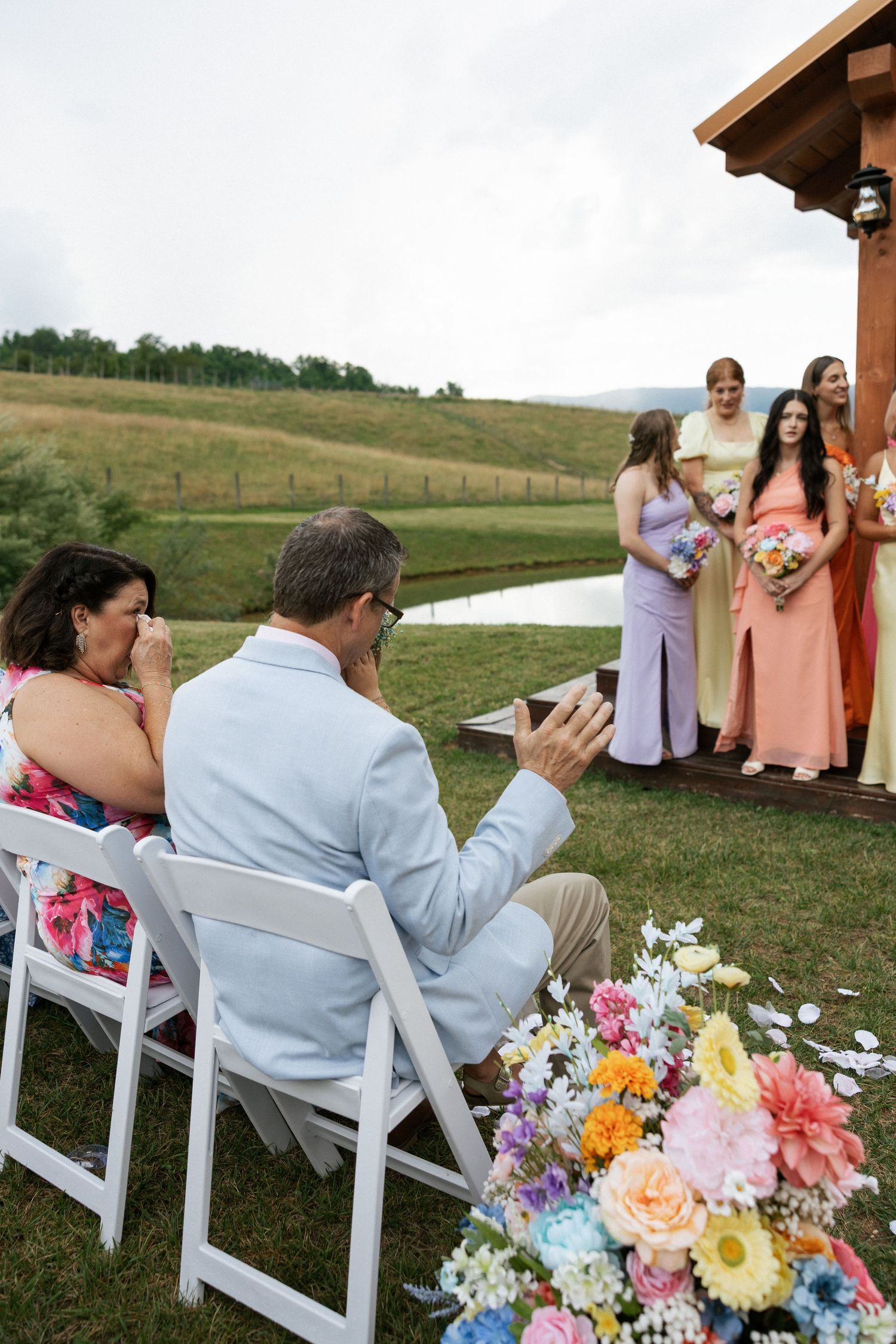 Wedding guests seated in white chairs with colorful floral arrangements at an outdoor ceremony.