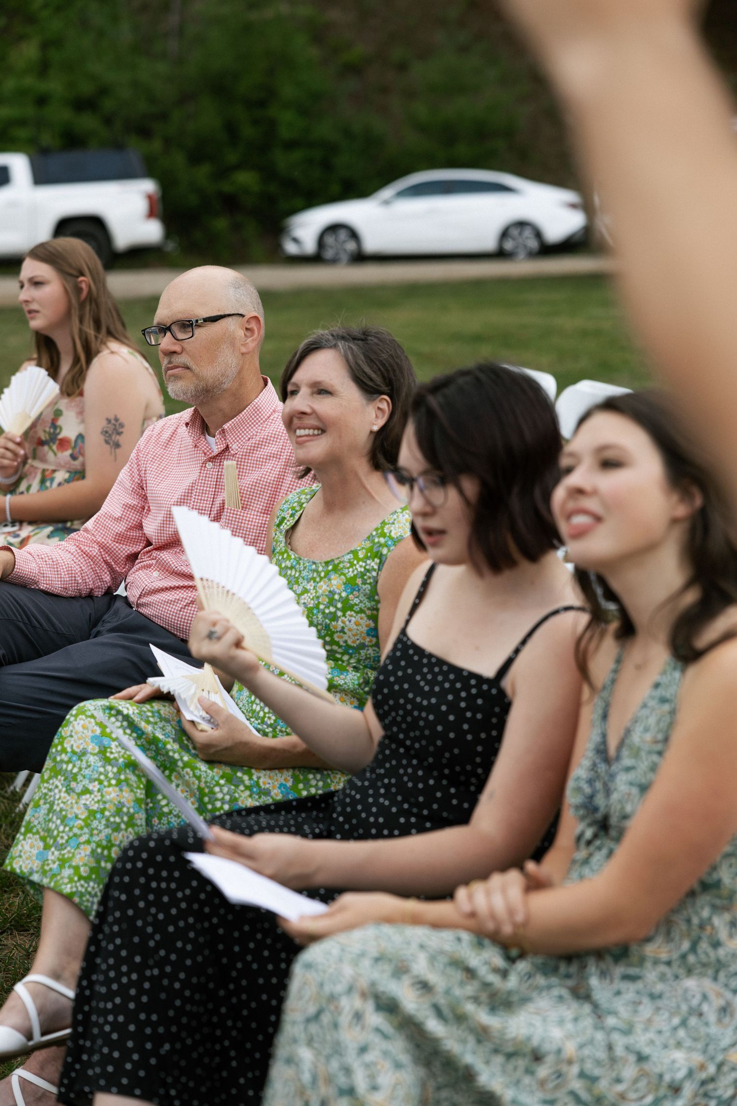Wedding guests seated outdoors holding paper fans during a summer ceremony.