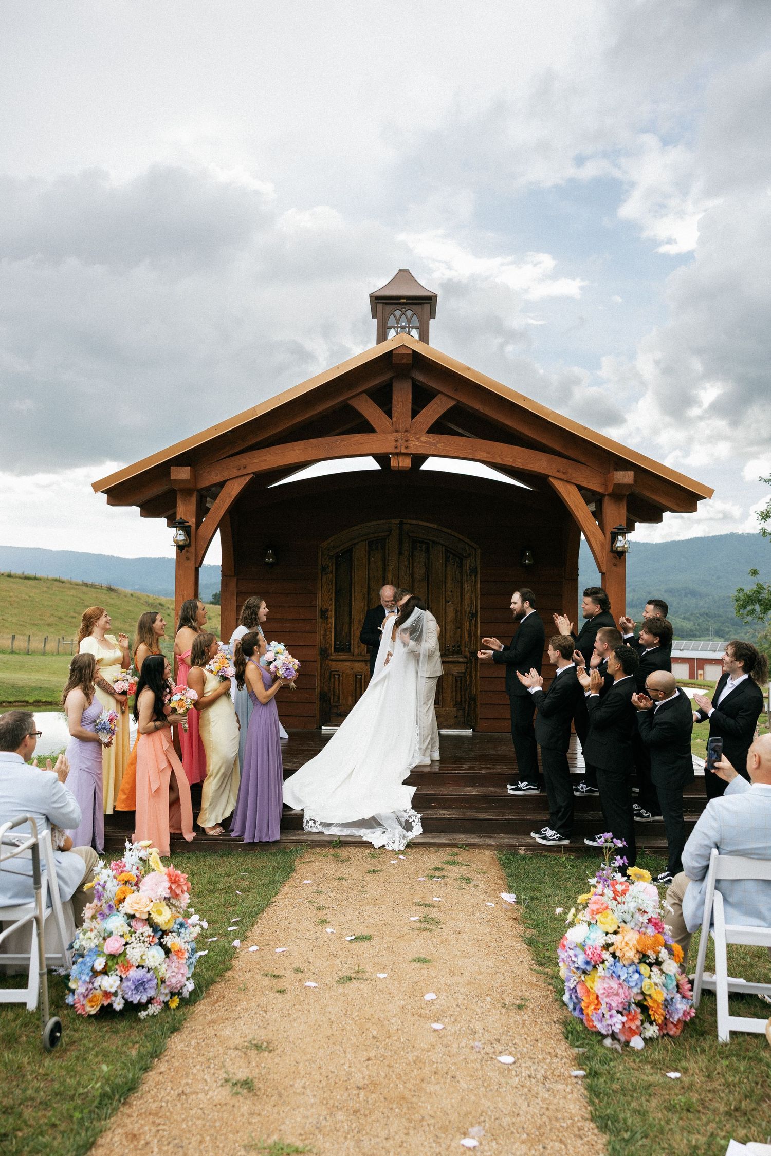 A rustic outdoor wedding ceremony with wooden pavilion, white chairs, and vibrant flower arrangements lining the aisle.