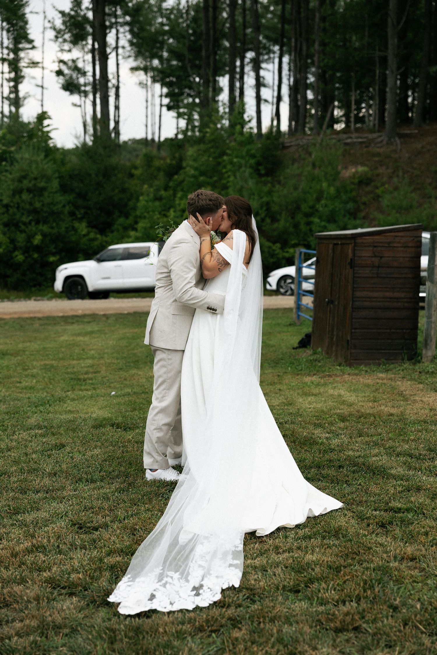 A couple shares their first kiss as newlyweds at an outdoor wedding with pine trees in the background.
