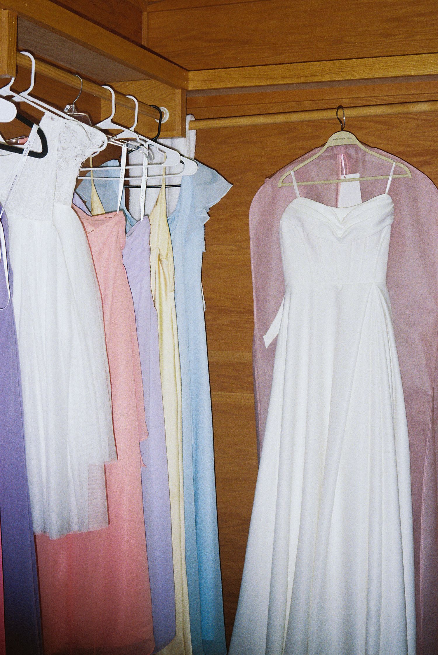 Pastel colored dresses and garments hanging on white hangers against a wooden wall display a soft color palette.