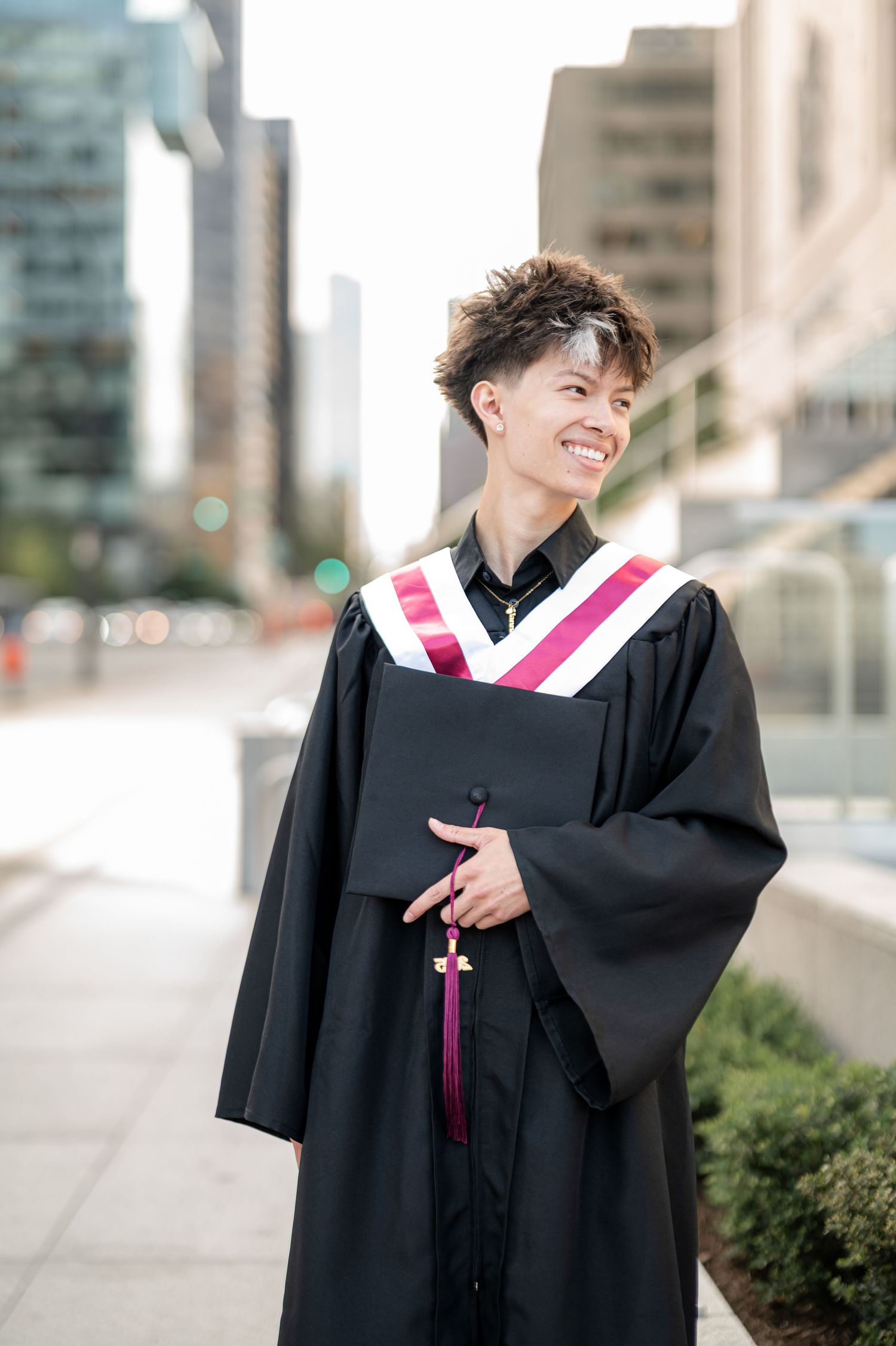 Graduate in black academic gown with pink stole smiling and posing for graduation photos on urban campus.