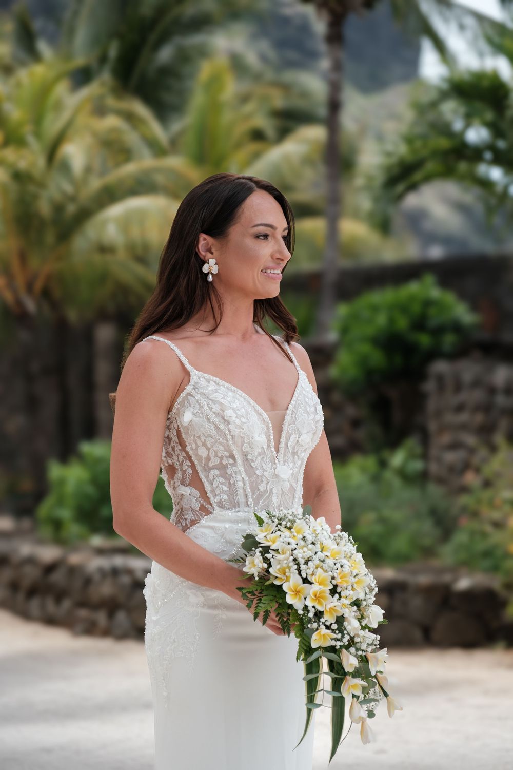 Elegant bride holding yellow and white cascading bouquet in tropical garden setting.