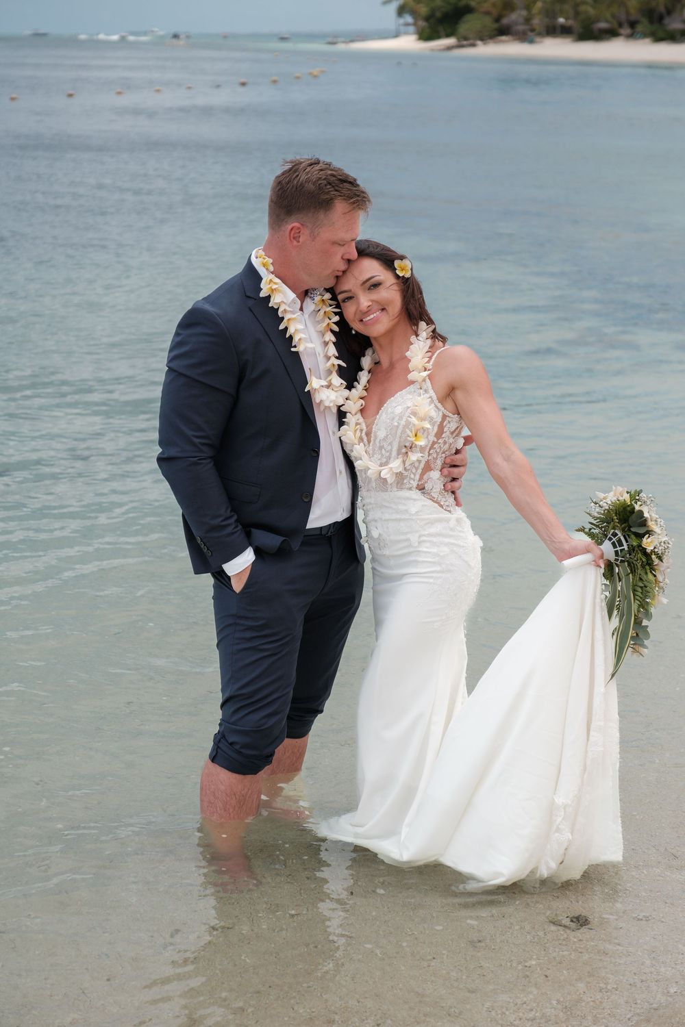Beach wedding couple wearing flower leis pose at waters edge in formal attire.
