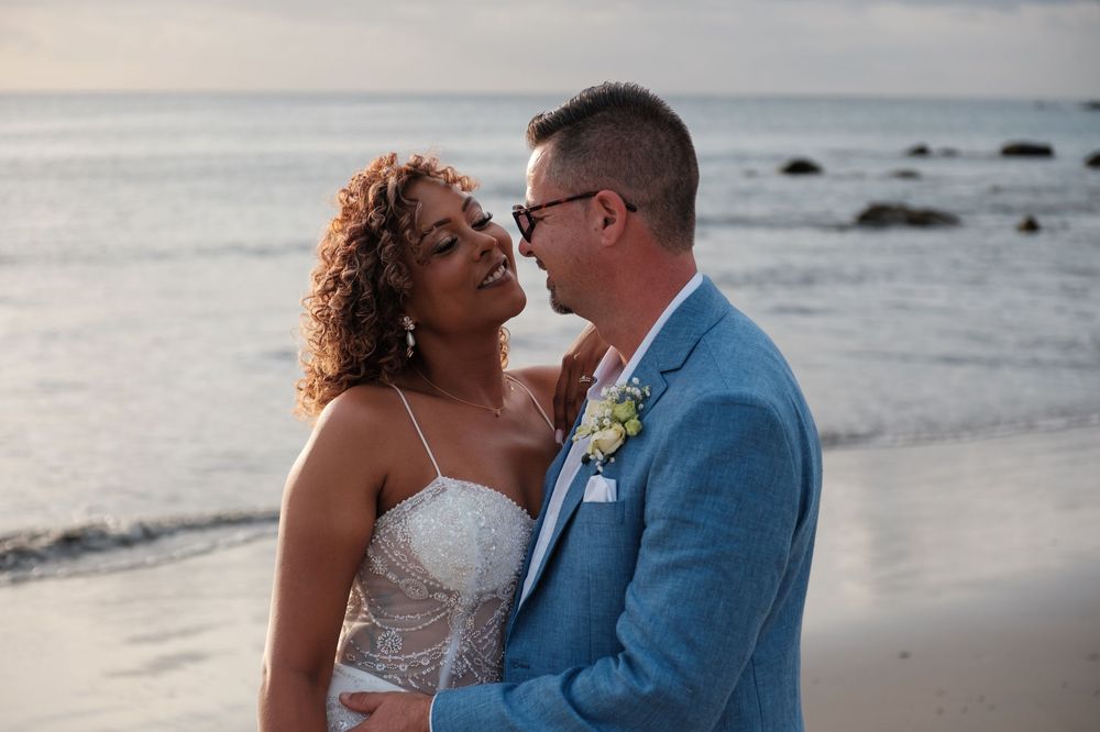 Intimate beach wedding portrait of couple in blue suit and white lace dress against ocean backdrop.