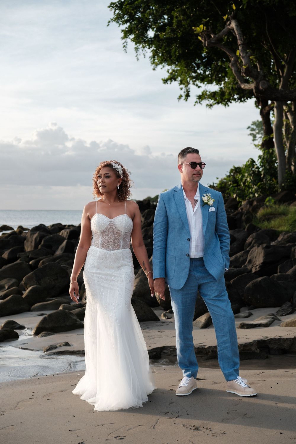 Newlyweds walking on tropical beach with bride in white gown and groom in powder blue suit.