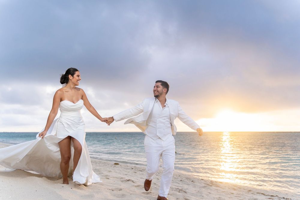 Joyful couple running on beach at sunset in flowing white wedding attire.