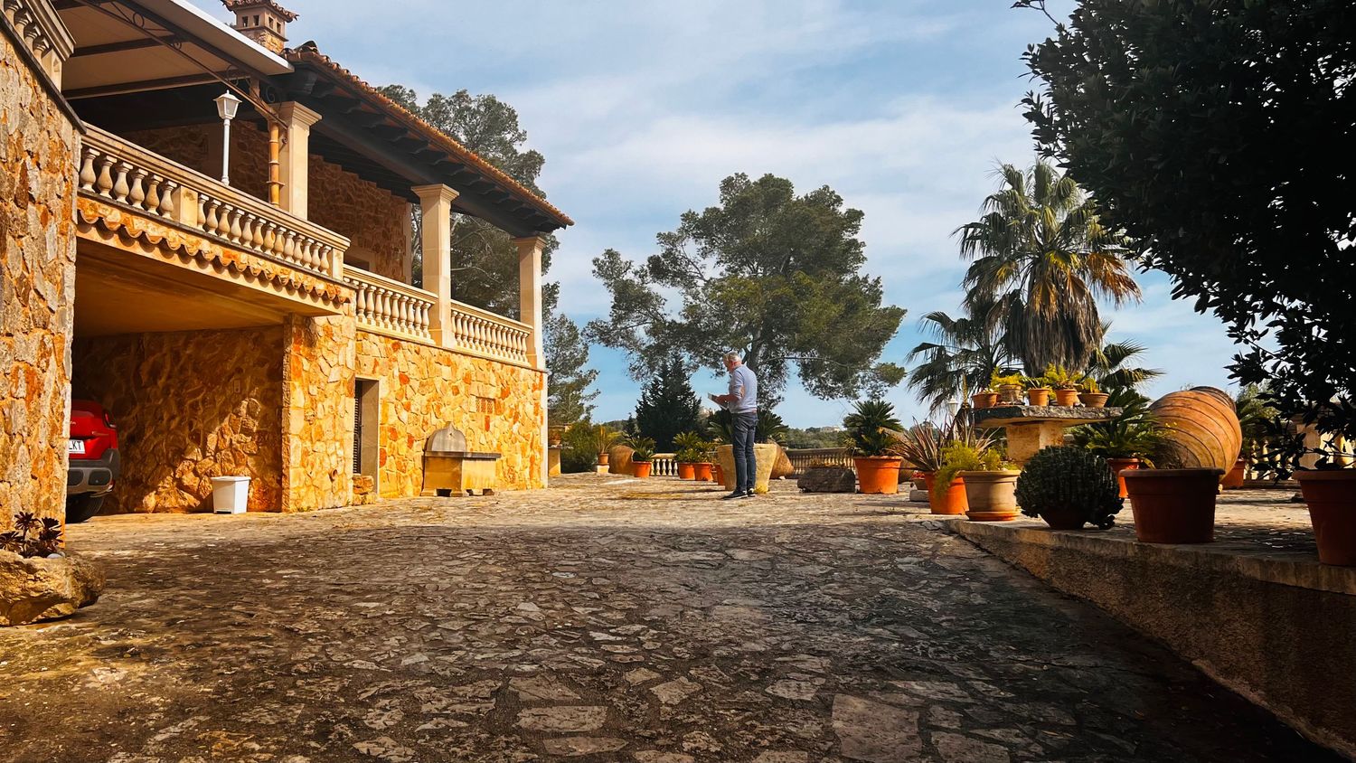 Rustic stone villa with cobblestone courtyard, palm trees, and terracotta pots bathed in warm golden sunlight.