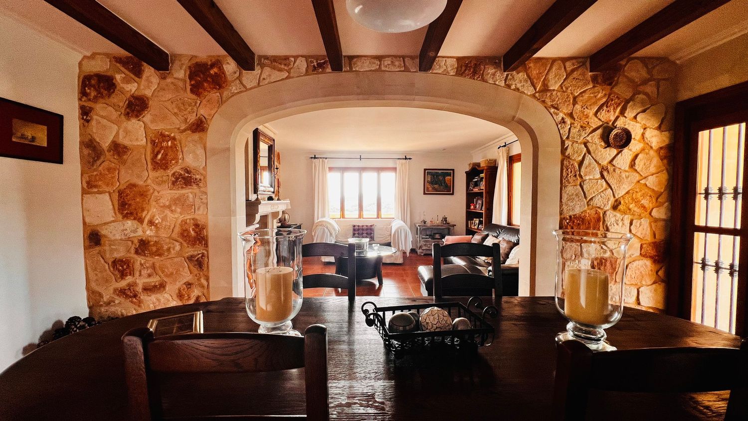 Rustic dining room with stone arch, wooden beams, leather chairs, and warm lighting leading to a cozy living area.