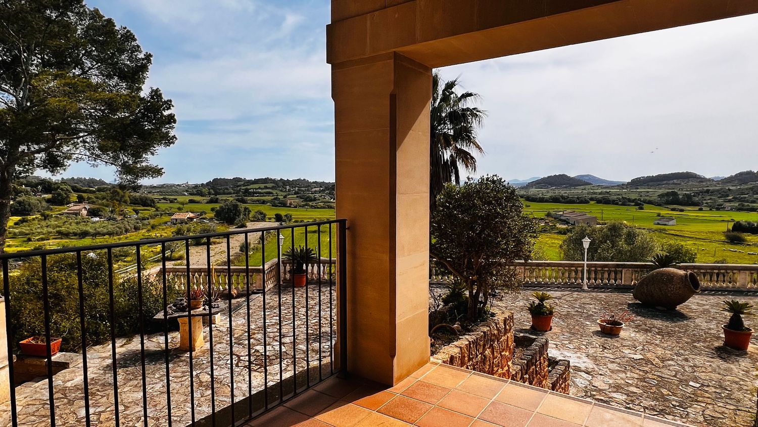 Scenic countryside view from a villa terrace with iron gate, palm tree, and green fields.