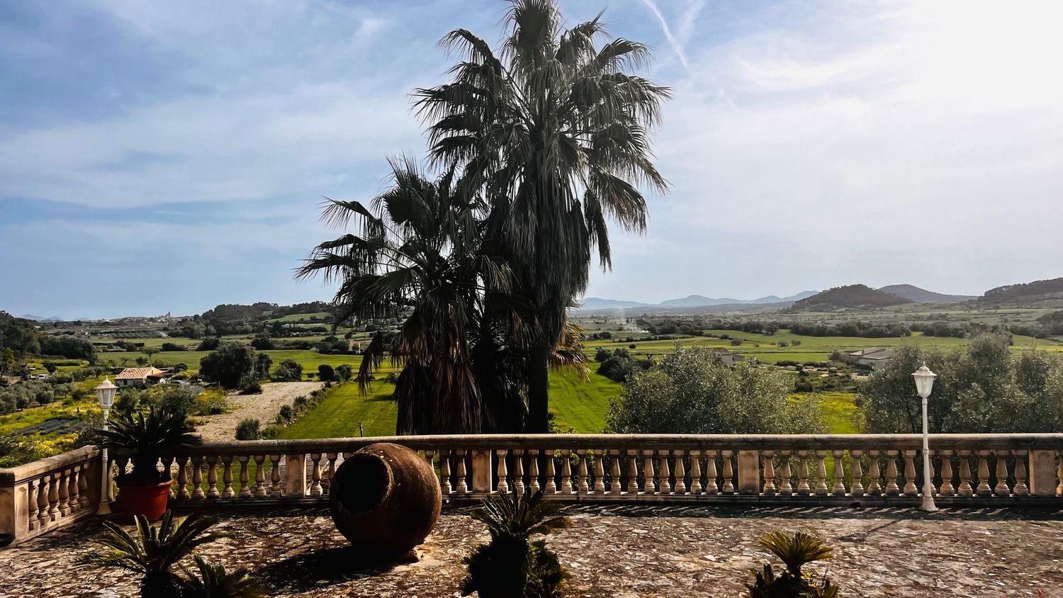 Panoramic view from a Mediterranean villa terrace with palm trees, lush gardens, and rolling hills under a blue sky.